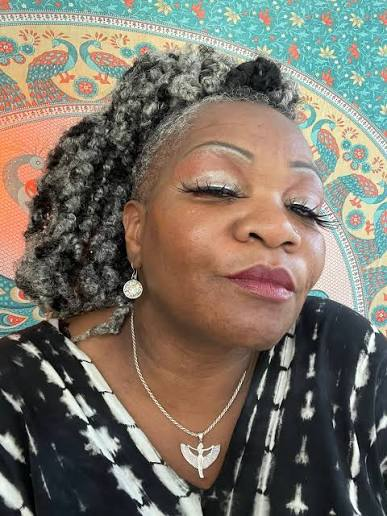 Colour portrait of Trina Meade smiling, silver locs and a winged pendant